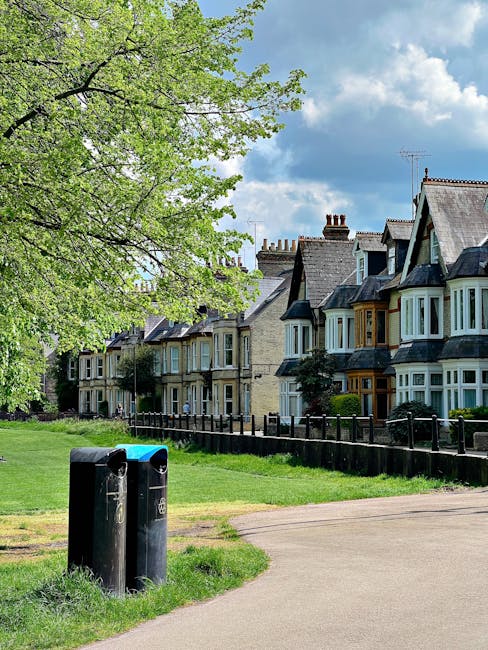 A row of historic terraced houses with brick facades, bay windows, and pitched roofs situated along a tree-lined street in a residential area. The houses have decorative architectural details and are painted in various muted tones, with some featuring contrasting window frames. In the foreground, there are two black waste bins placed on a paved footpath next to a grassy verge. A large, leafy green tree extends across the top left corner of the image, with fresh spring foliage. The sky above is partly cloudy with patches of blue, indicating a clear daytime setting. The scene suggests a peaceful, well-maintained neighbourhood where private waste collection services may be used, supported by local rubbish removal companies like House Clearance Sutton, instead of municipal services. The overall environment emphasizes community residential living with a focus on tidiness and curb appeal.