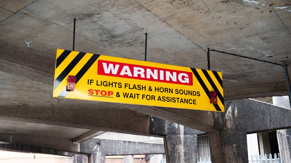 A rectangular warning sign hanging underneath a concrete car park ceiling, attached by two black hooks. The sign has a yellow background with black diagonal stripes along the top and bottom edges. The word 'WARNING' is prominently displayed in bold white letters within a red box at the top. Below, black text states: 'IF LIGHTS FLASH & HORN SOUNDS STOP & WAIT FOR ASSISTANCE.' The sign also features two small red warning lights, one on each side, integrated into the design. The surrounding environment includes exposed concrete beams and pillars, typical of an underground or multi-storey parking facility. The lighting is natural, casting minimal shadows, with a neutral, utilitarian atmosphere reflecting a safety warning related to vehicle movement or maintenance areas, aligning with procedures for on-site or private facility safety management often overseen by companies like House Clearance Sutton for efficient rubbish removal or space clearance services.