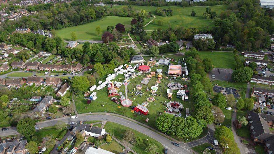 An aerial view of a community fairground set up in a grassy park area surrounded by residential housing and lush green trees. The fairground features a variety of attractions, including a red and white striped tent, colorful amusement rides such as a small Ferris wheel, and several food stalls with striped canopies. Numerous caravans and trailers are parked in clusters around the site, some appearing to serve as vendor booths or storage units. The area is bordered by a curved road with cars parked along the roadside, and a dense line of mature trees separates the park from adjacent residential streets. In the background, open green fields and wooded areas extend beyond the fairground, highlighting the contrast between the supervised event space and the surrounding natural landscape. The scene is illuminated by daylight with clear weather, indicating a busy, organized outdoor event, possibly scheduled for rubbish clearance following the event period, as managed by House Clearance Sutton's rubbish removal services.