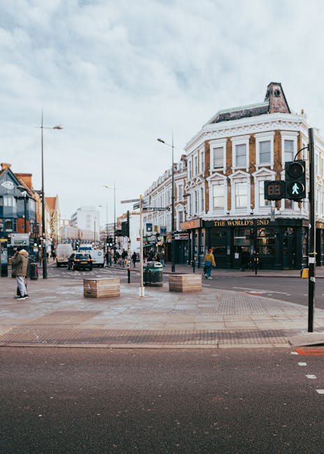 A busy street scene on Sutton High Street featuring a prominent three-story corner building with a curved façade, white decorative architectural details, and large windows, housing a pub named 'The World's End' on the ground level. Surrounding the building are pedestrians walking along the wide paved pedestrian area, some crossing at a traffic light that displays a green pedestrian signal. Several wooden planter boxes with no plants are positioned on the pavement, and a variety of vehicles, including cars and vans, are visible on the street, indicating typical urban activity. The background includes additional shops and modern buildings under a partly cloudy sky, with streetlights and traffic signage present, reflecting a lively town centre environment. This scene relates to municipal or private rubbish removal services operating within the urban setting, highlighting everyday waste management needs in high street areas, supported by companies like House Clearance Sutton or similar providers specializing in alternative waste handling solutions.