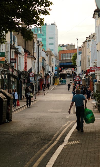 A busy street scene on Sutton High Street featuring a prominent three-story corner building with a curved façade, white decorative architectural details, and large windows, housing a pub named 'The World's End' on the ground level. Surrounding the building are pedestrians walking along the wide paved pedestrian area, some crossing at a traffic light that displays a green pedestrian signal. Several wooden planter boxes with no plants are positioned on the pavement, and a variety of vehicles, including cars and vans, are visible on the street, indicating typical urban activity. The background includes additional shops and modern buildings under a partly cloudy sky, with streetlights and traffic signage present, reflecting a lively town centre environment. This scene relates to municipal or private rubbish removal services operating within the urban setting, highlighting everyday waste management needs in high street areas, supported by companies like House Clearance Sutton or similar providers specializing in alternative waste handling solutions.